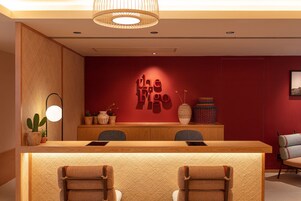 Un área de recepción con un mostrador de madera, dos sillas, una pared roja con un logotipo, una lámpara colgante y objetos decorativos.