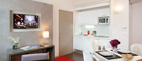 A modern hotel room with a dining area, a TV mounted on the wall, a small desk with a lamp, and a red carpet.