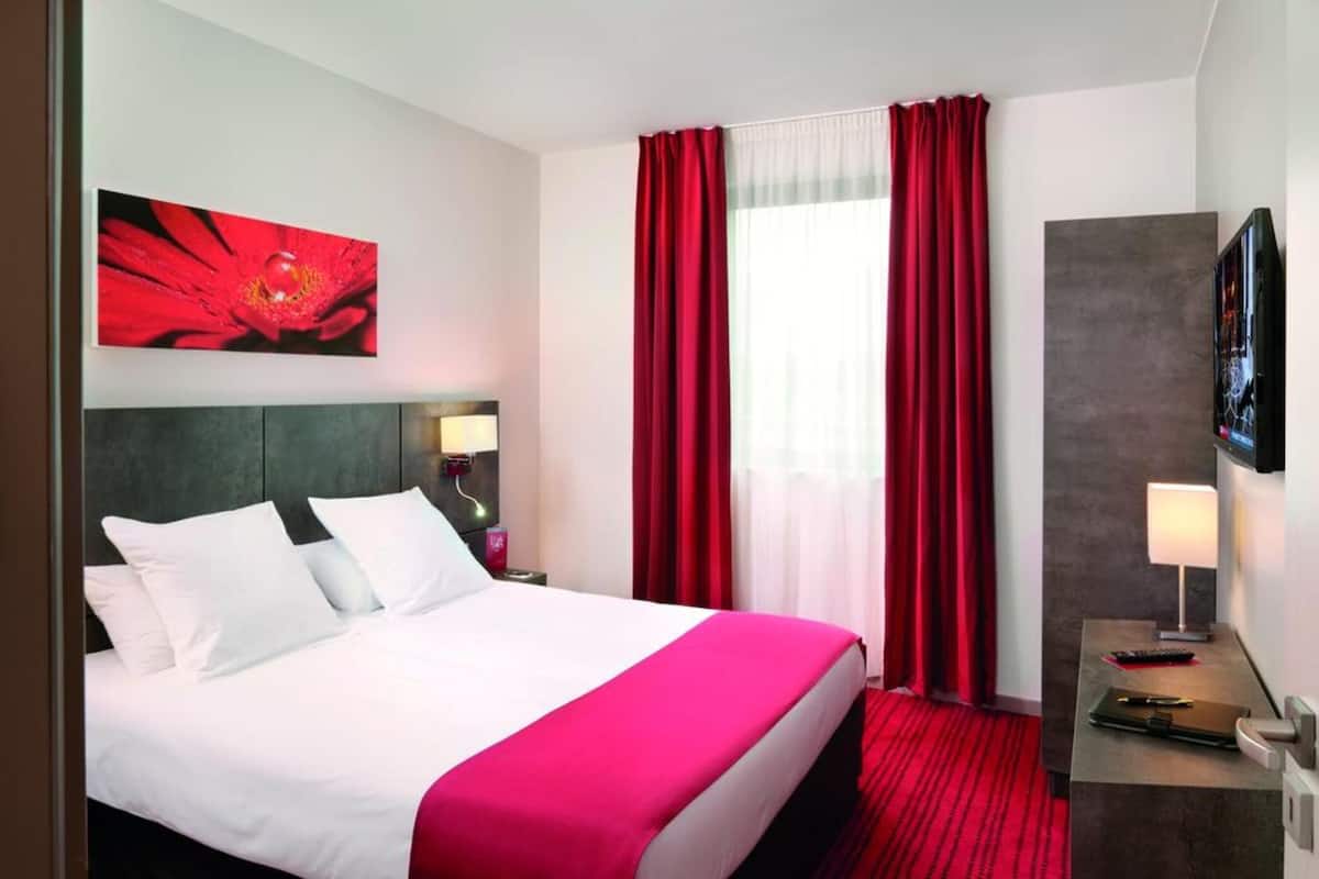 Hotellihuone, jossa on sänky, kirjoituspöytä lampun kanssa, televisio ja punainen verho.