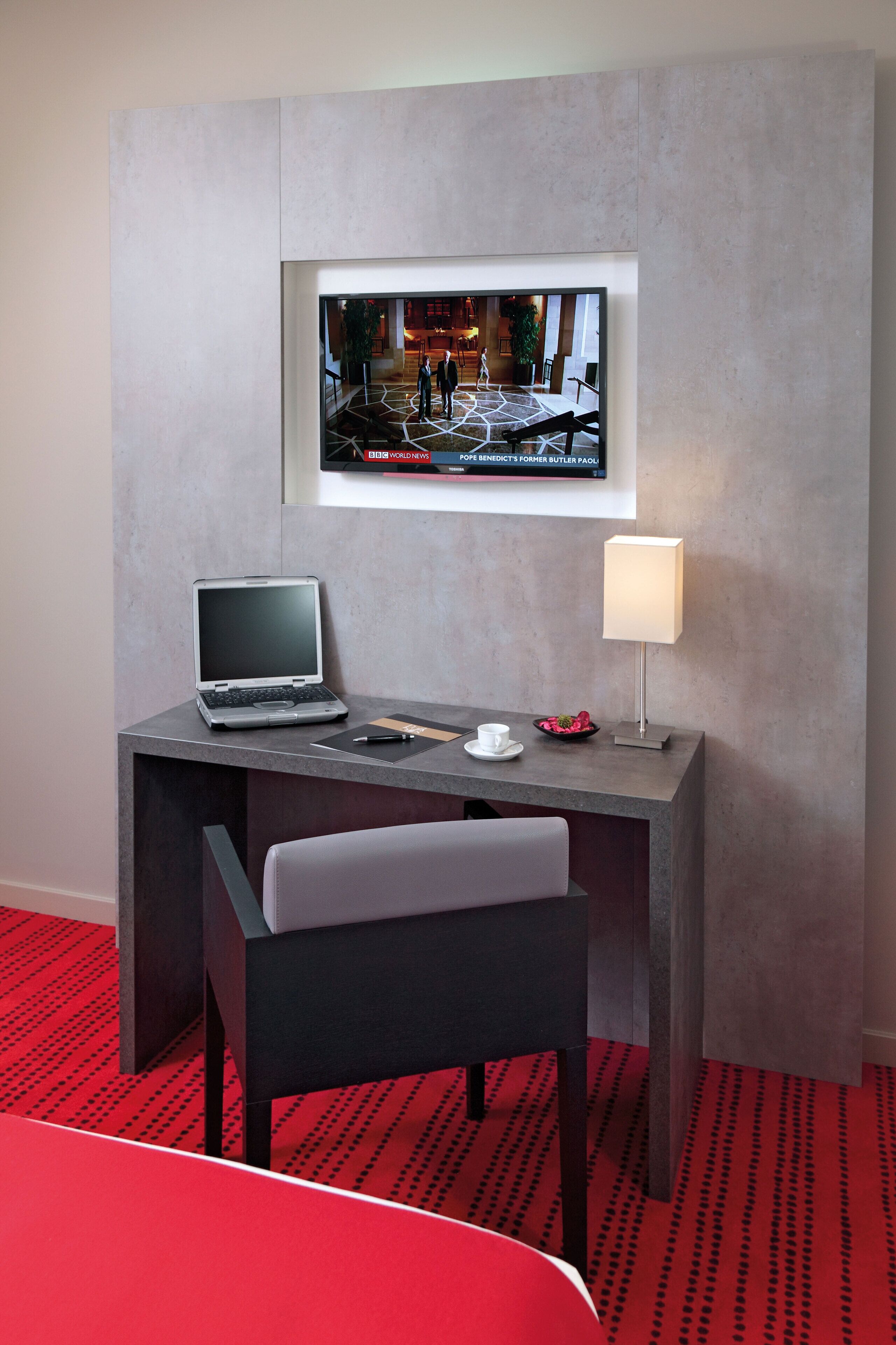 A modern hotel room with a desk, laptop, chair, and a television mounted on the wall displaying a news channel.