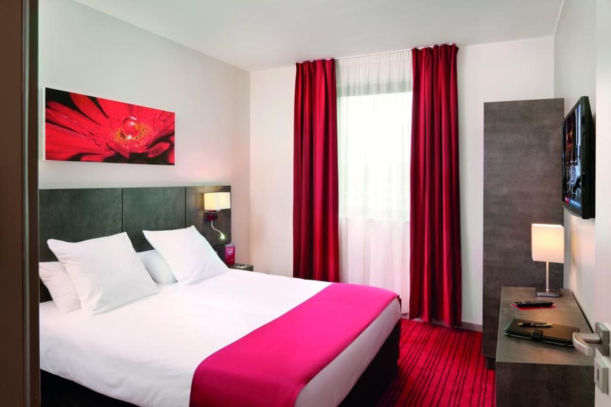 A hotel room with a bed, a desk with a lamp, a TV, and a red curtain.