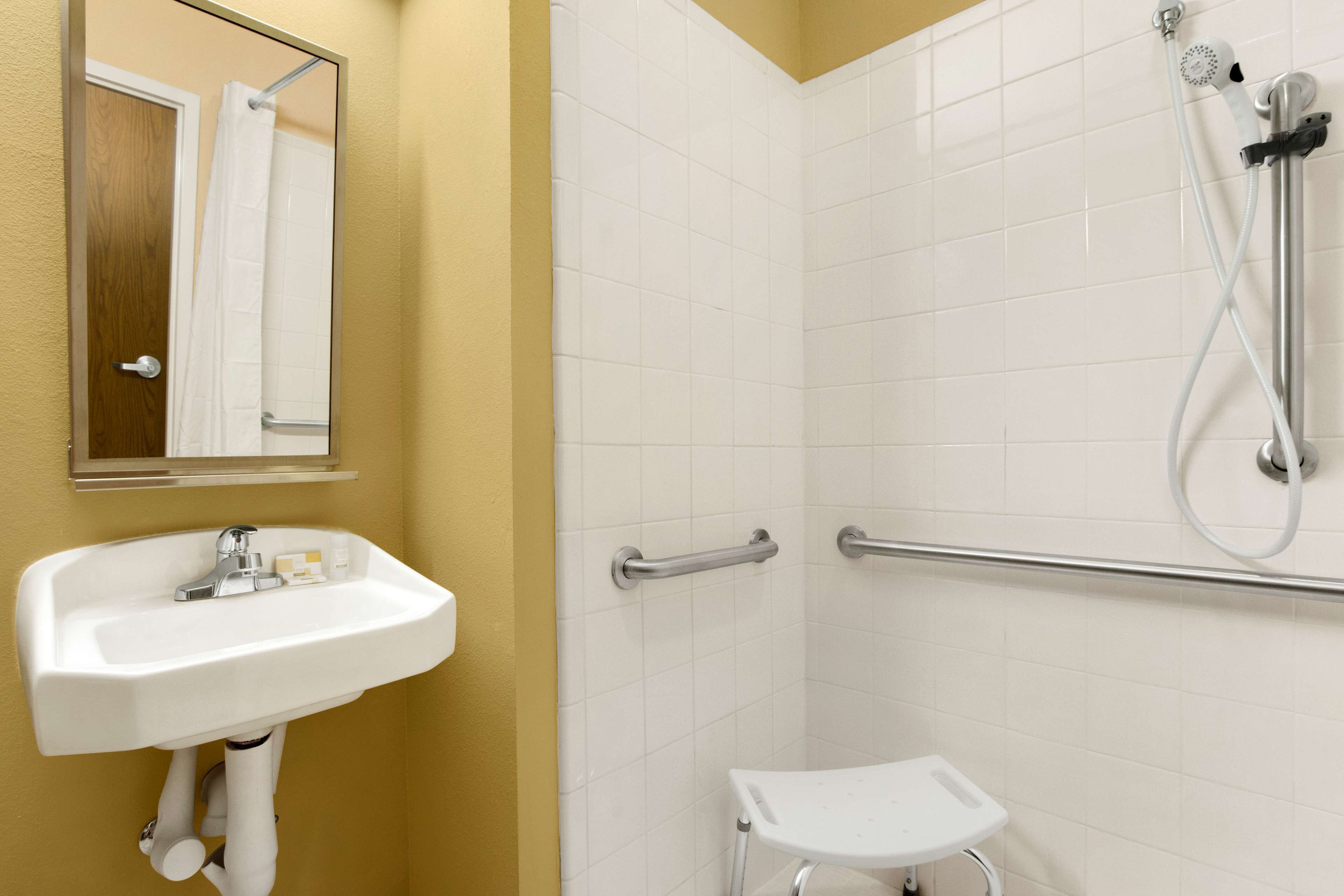 Un baño con lavamanos blanco, un espejo, ducha con cabezal de ducha manual y una barra de apoyo.