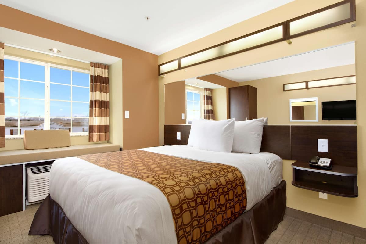 Ein Hotelzimmer mit einem großen Bett, einem Fenster mit Vorhängen, einem Nachttisch mit Telefon und einem an der Wand montierten Flachbildfernseher.