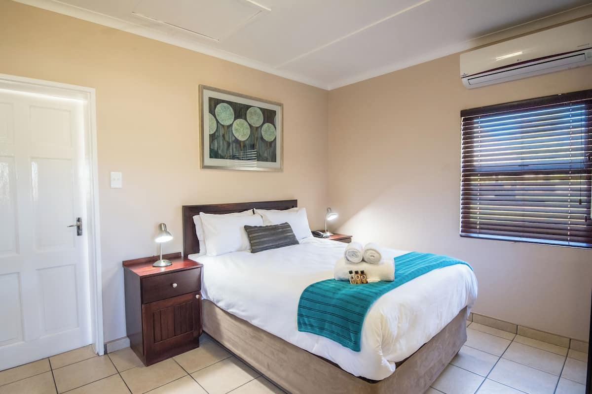 Une chambre à coucher bien rangée, avec un lit, des tables de chevet, une œuvre d’art encadrée, un appareil de climatisation et une fenêtre avec des stores.