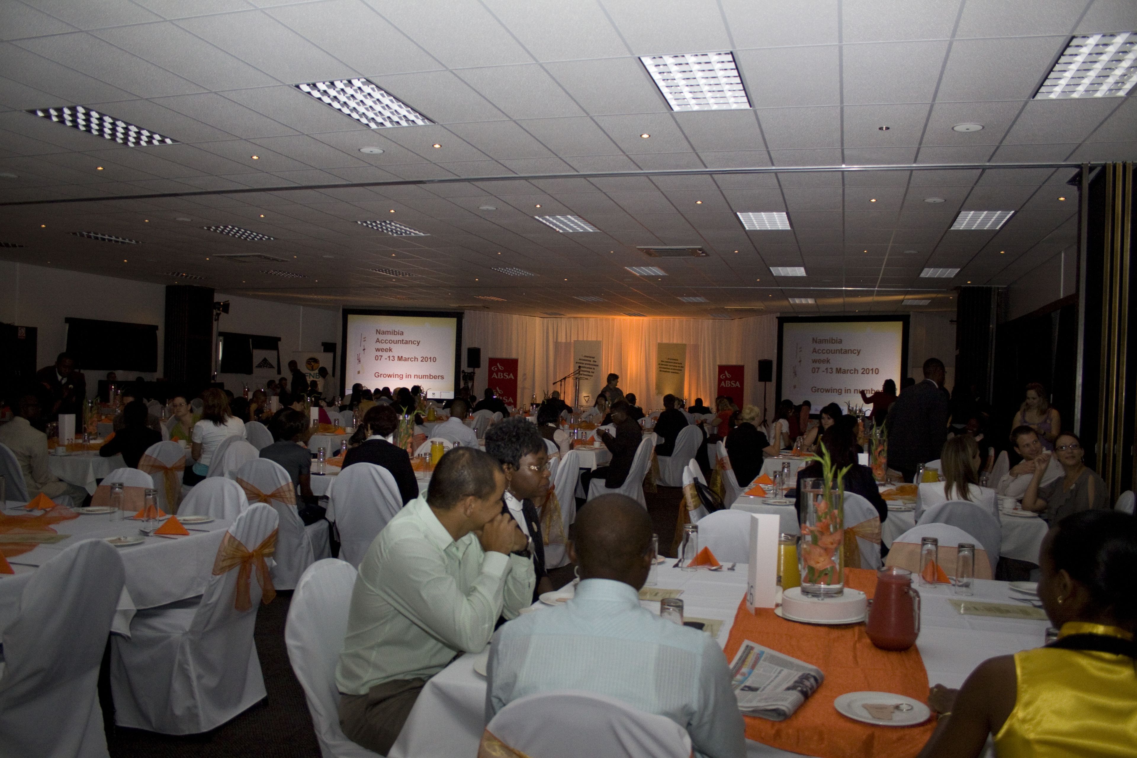 Un salón de conferencias con varias mesas dispuestas para un evento formal, con tarjetas de identificación y pantallas de presentación.