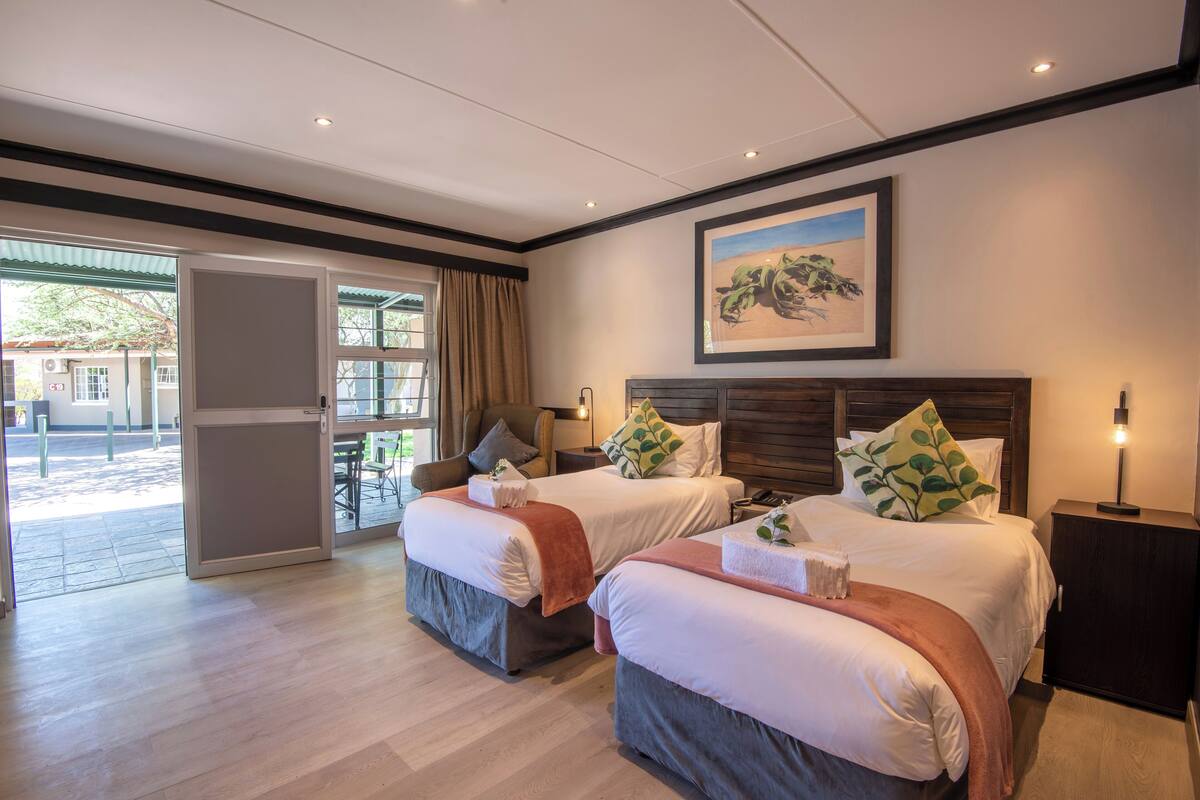 Une chambre d’hôtel avec deux lits, une grande fenêtre, une porte donnant sur l’extérieur et un tableau accroché au mur.