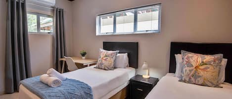 A hotel room with two beds, a desk, a chair, a window with blinds, and a ceiling-mounted air conditioner.