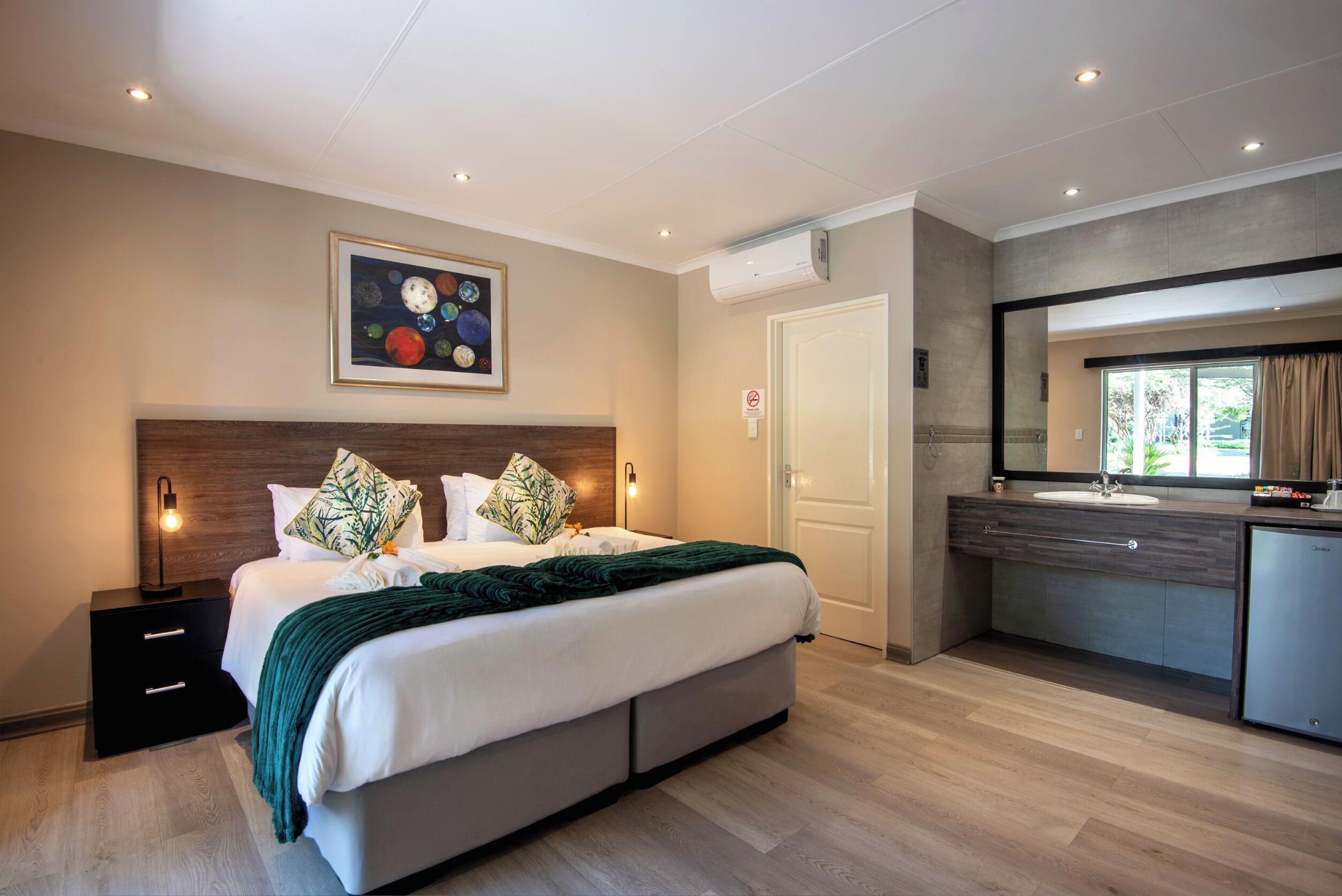 A modern hotel room with a large bed, a bedside table, a built-in desk, a bathroom with a sink, and a large window.