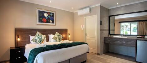 A modern hotel room with a large bed, a bedside table, a built-in desk, a bathroom with a sink, and a large window.