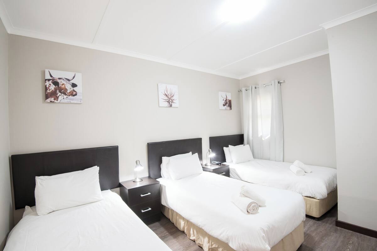 Une chambre d’hôtel avec trois lits, une table de chevet avec une lampe et des œuvres d’art accrochées aux murs.