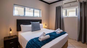A neatly made bed with a blue blanket, a bedside table with a lamp, and a window with curtains.
