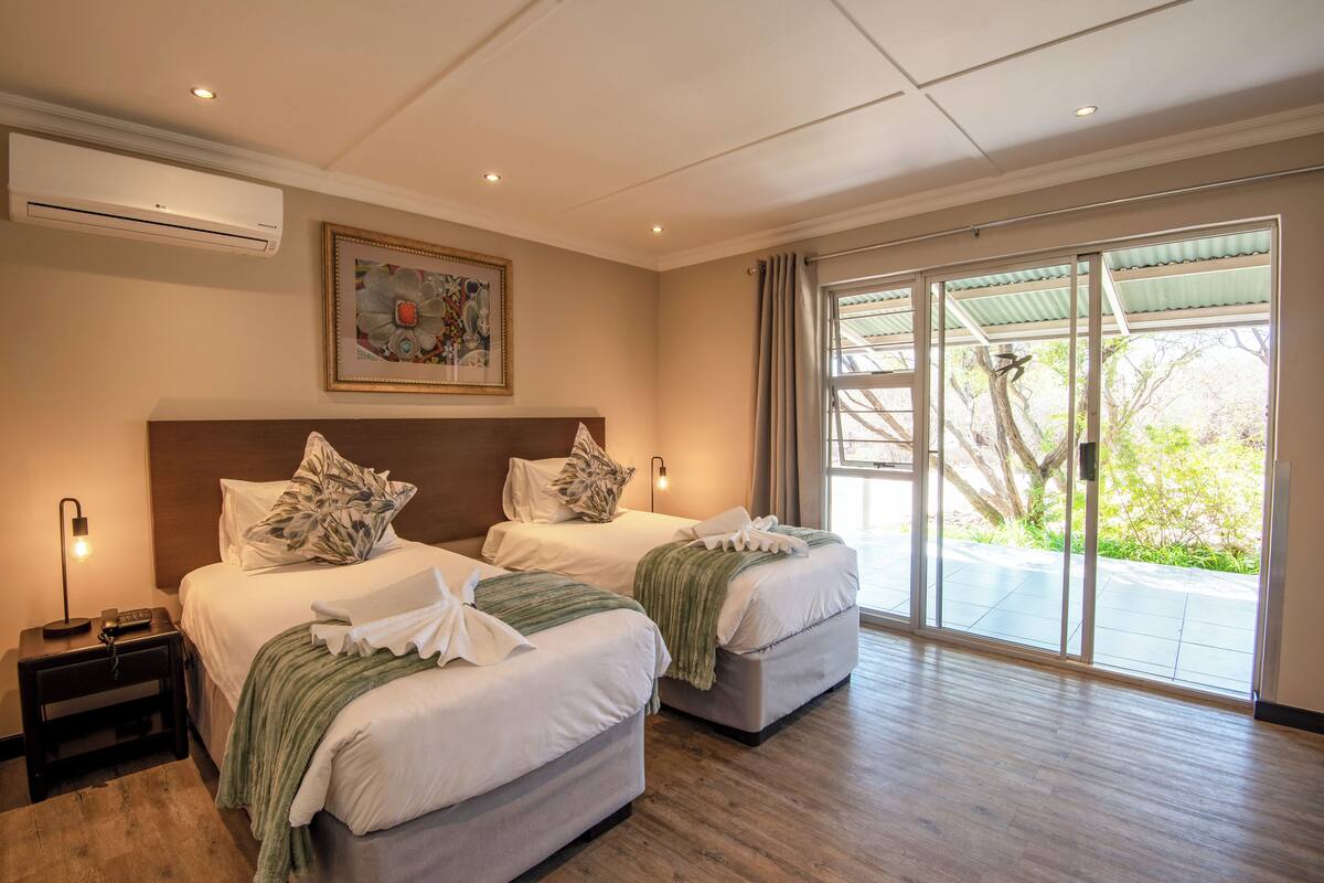 Une chambre d’hôtel avec deux lits, une table de chevet, un appareil de climatisation, un tableau et une vue sur l’extérieur.