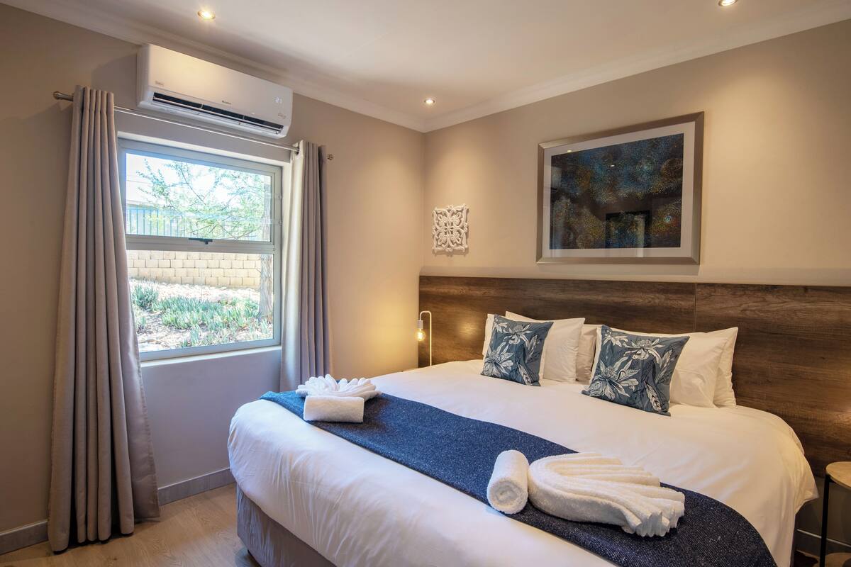 Une chambre d’hôtel avec un grand lit, une fenêtre avec des rideaux, un appareil de climatisation, une œuvre d’art accrochée au mur et une table de chevet.