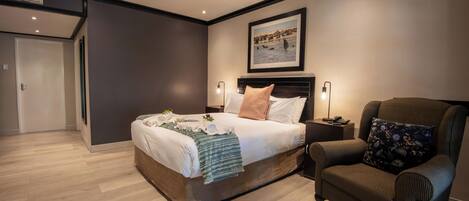 A modern bedroom with a large bed, a chair, a framed picture on the wall, and a door in the background.