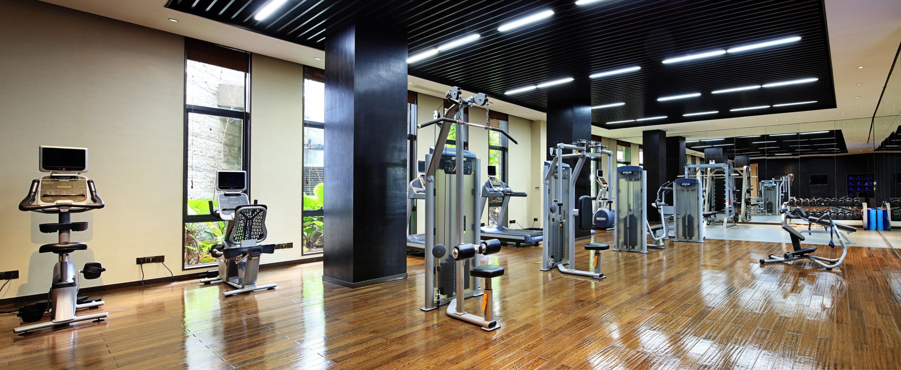 A gym with various exercise equipment, large windows, and a wooden floor.