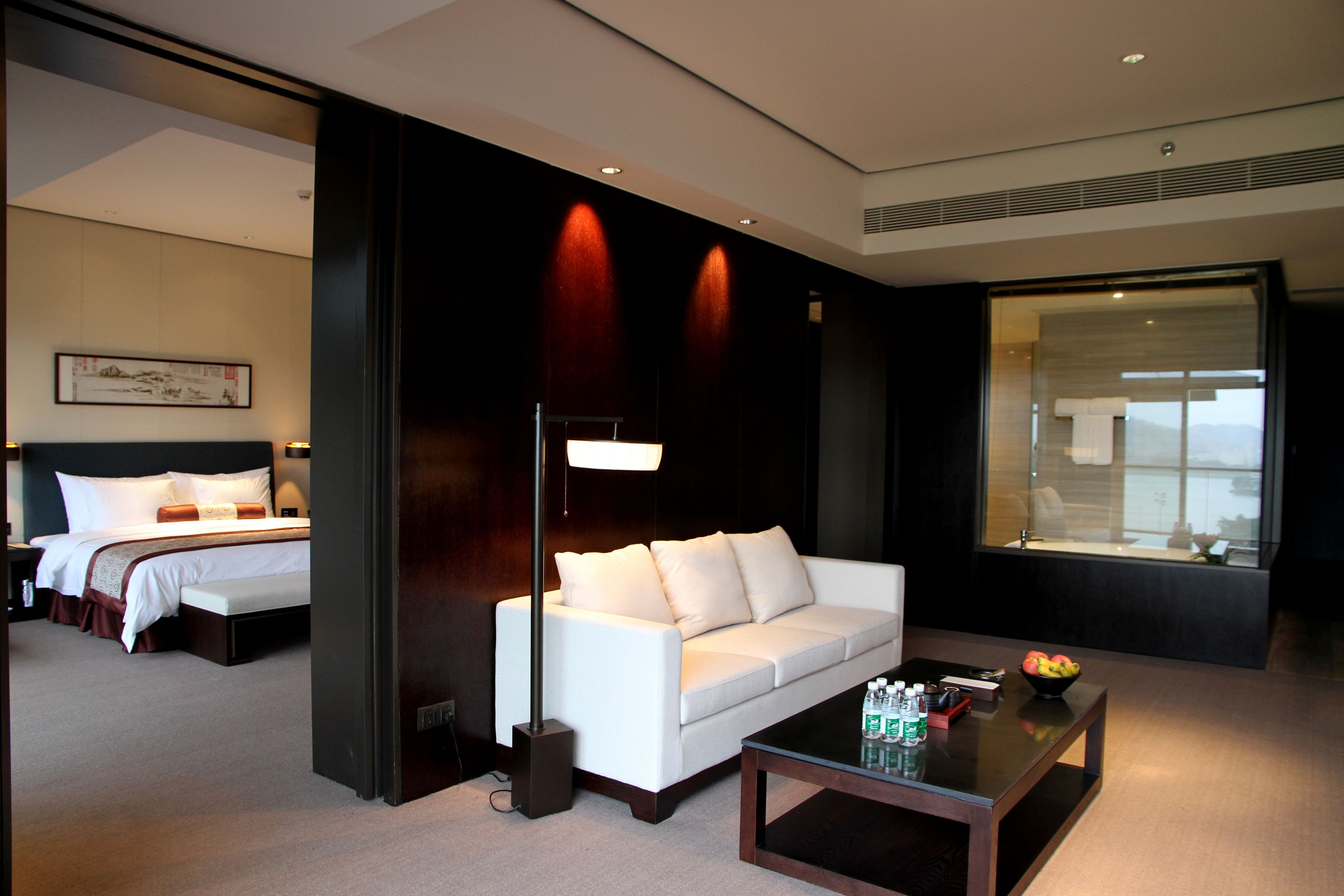 A hotel room with a bed, a white sofa, a coffee table with fruit and bottles, and a large window.