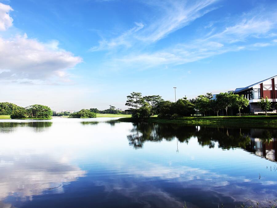 Un lago sereno refleja el cielo y los árboles, rodeado de follaje y un edificio con una fachada distintiva roja y blanca.