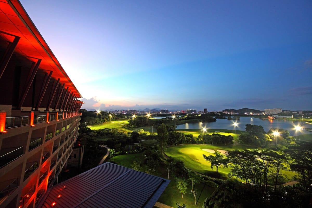 A modern building with a red-lit facade overlooking a well-lit golf course and a body of water at dusk.
