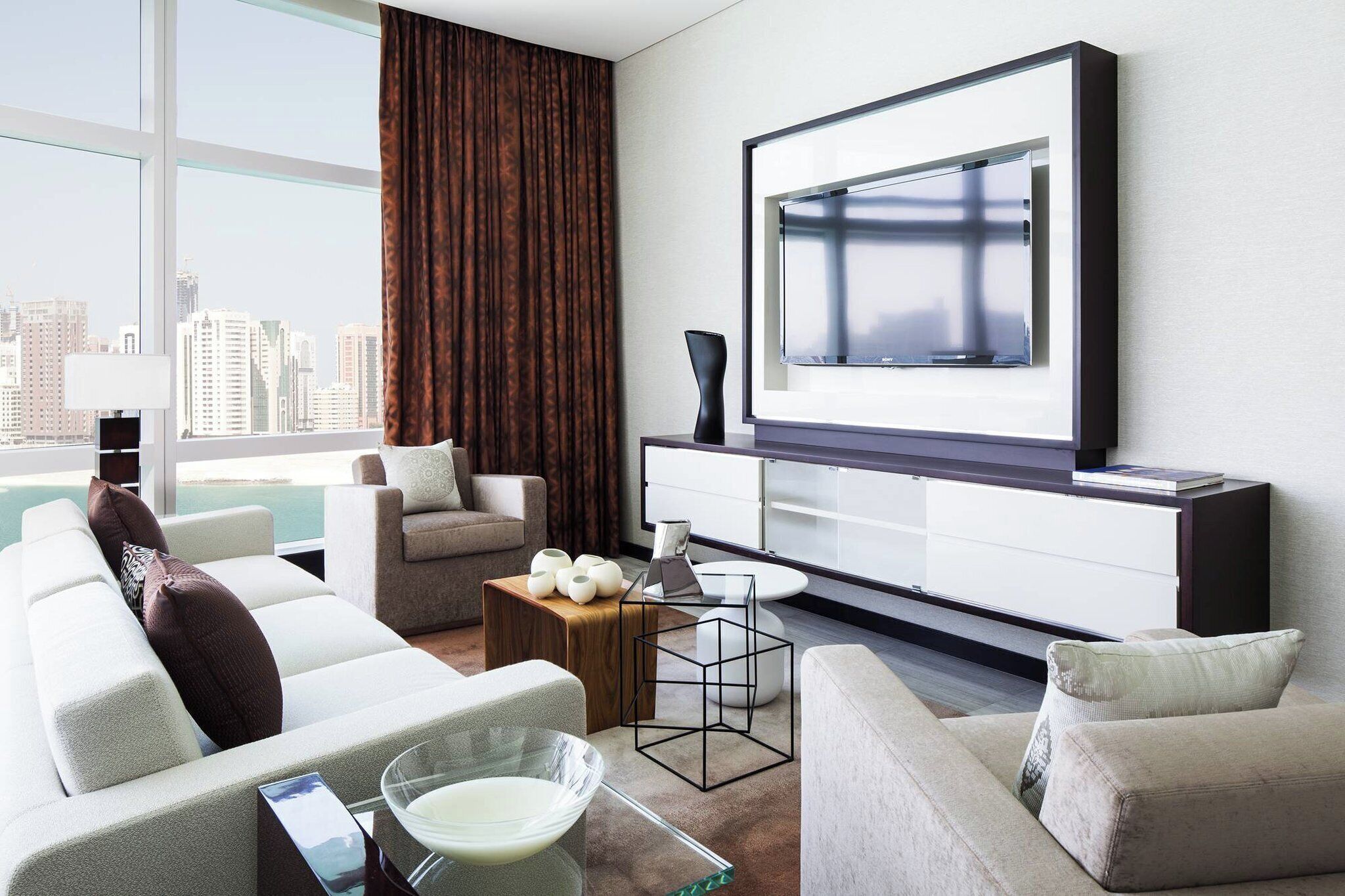 A modern living room with a large TV, comfortable seating, a coffee table, and a view of the city.