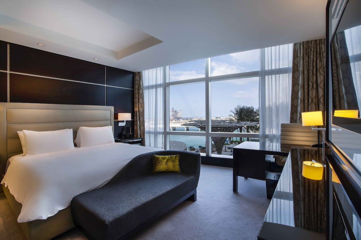 Une chambre d’hôtel moderne dotée d’un grand lit, d’un canapé, d’un bureau et offrant une vue sur le paysage urbain grâce à de grandes fenêtres.
