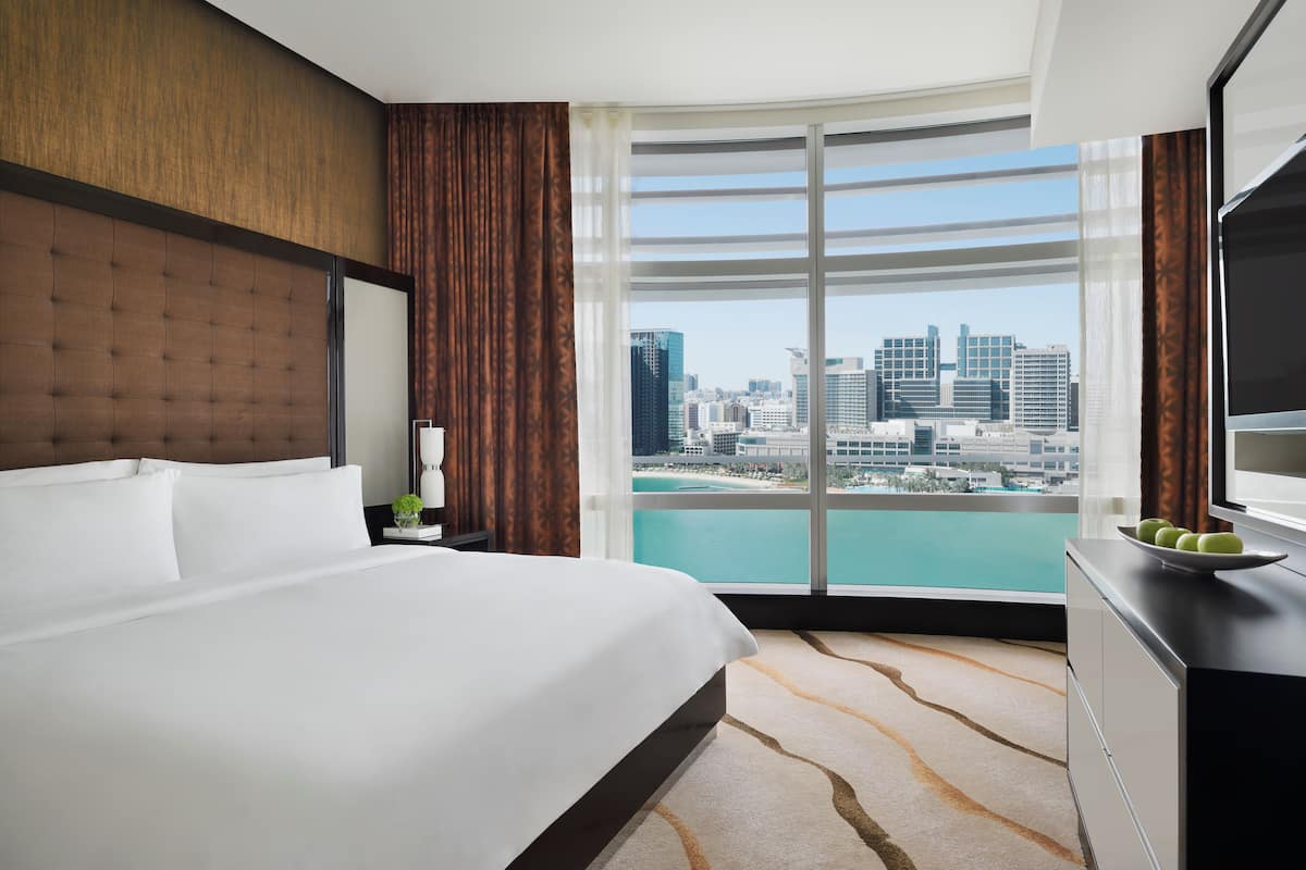 Une chambre d’hôtel avec un grand lit, une table de chevet, une commode et une vue sur les toits de la ville et sur l’eau.
