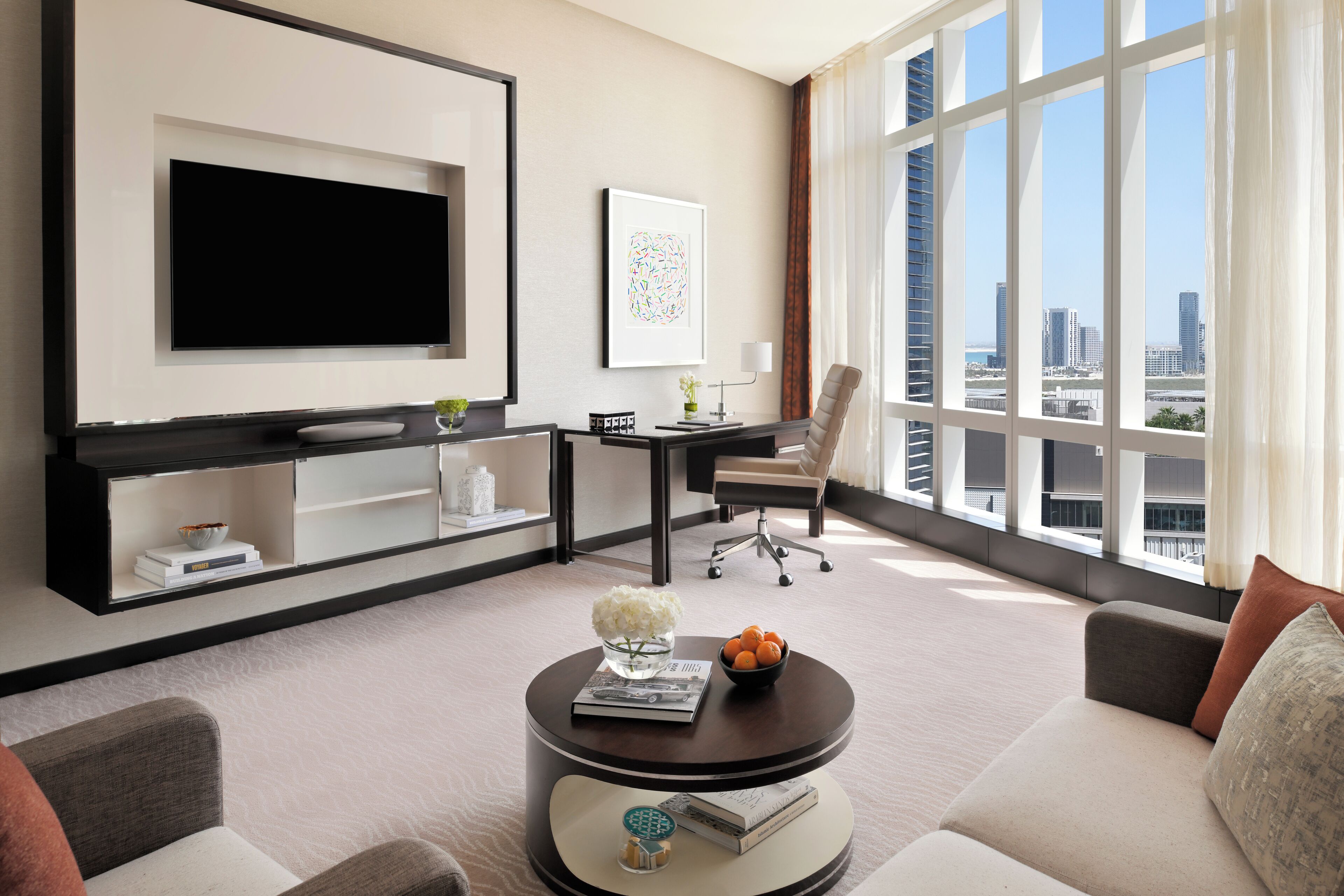 A modern hotel room with a large TV, a desk with a chair, a sofa, and a view of the city.