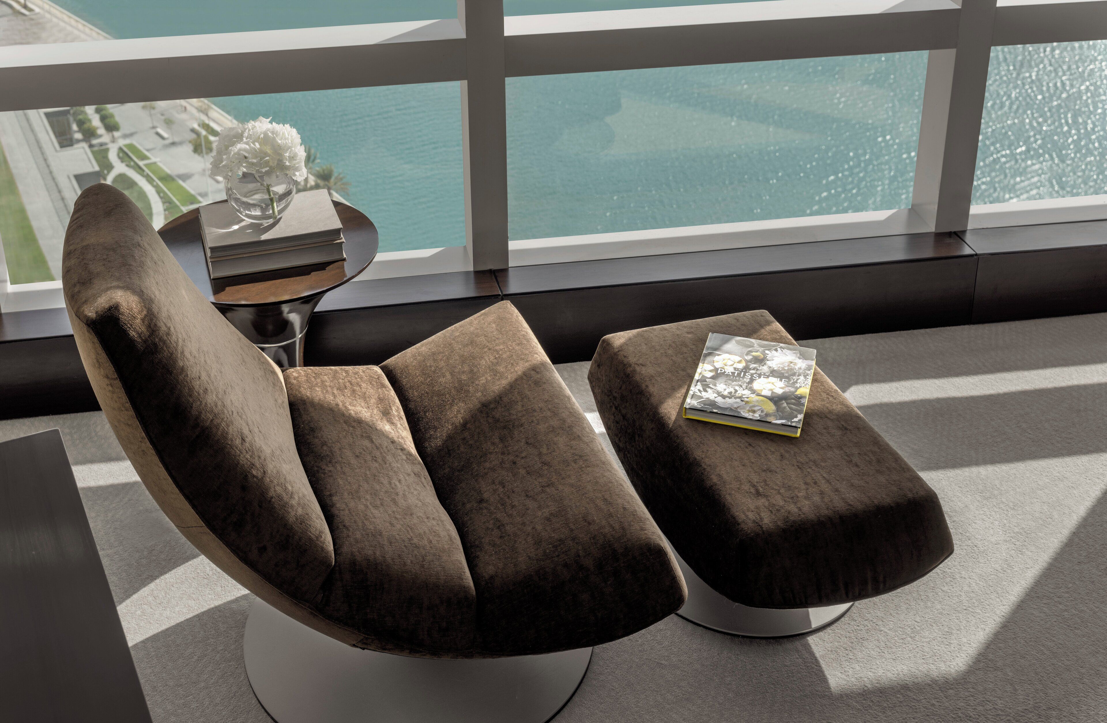 Una habitación moderna con una silla acolchada y un reposapiés, una mesita con un jarrón y libros, y vista al mar a través de amplias ventanas.