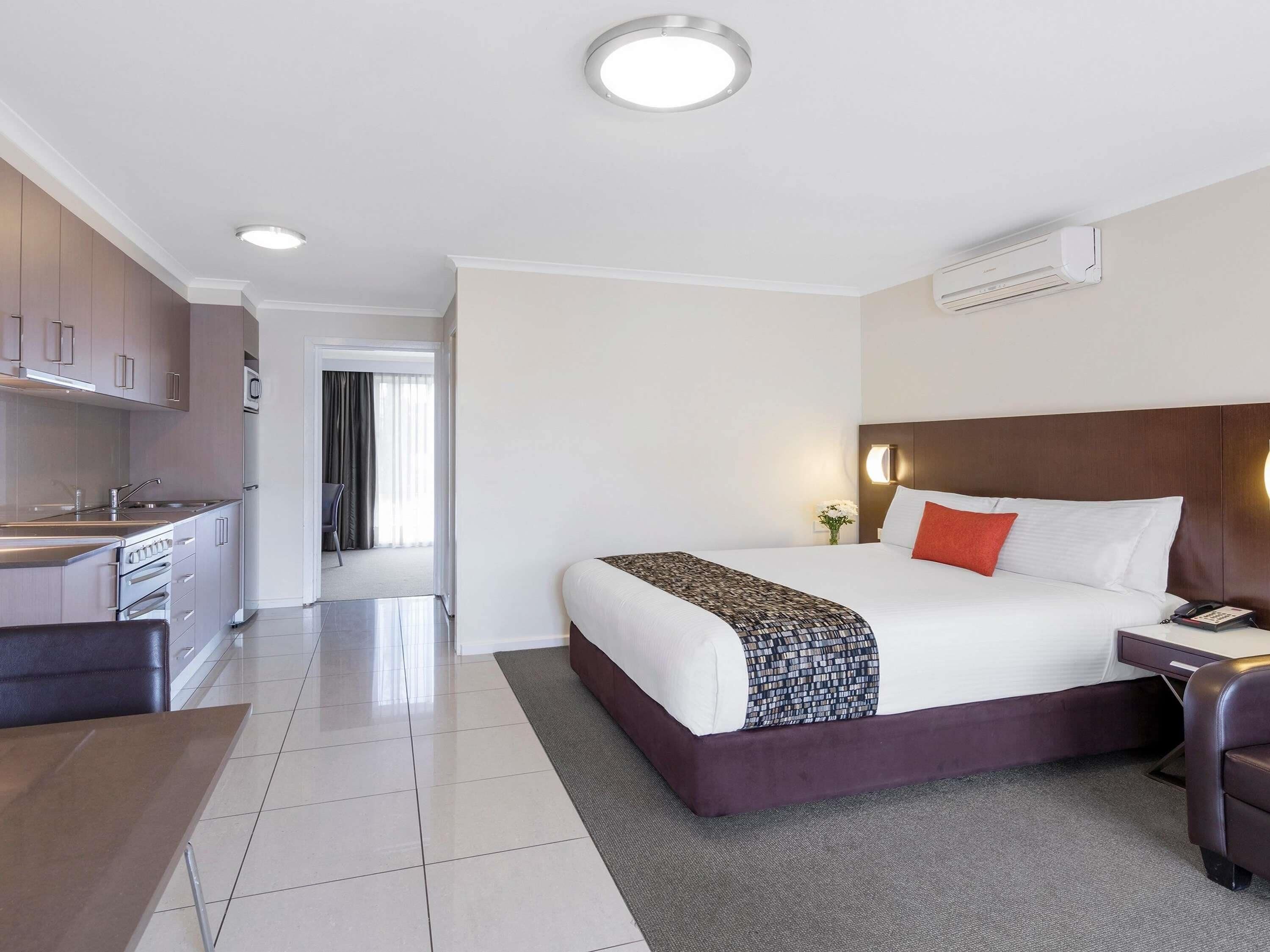 Une chambre d’hôtel comprenant un lit, une table de chevet, une lampe et donnant sur un coin cuisine.