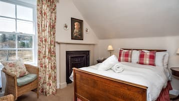 A bedroom with a large bed, two bedside tables with lamps, a wicker chair, a fireplace, and a view of the outdoors.