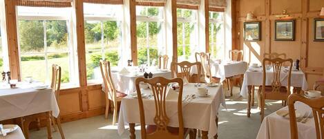 A dining area with multiple tables and chairs, large windows with curtains, and framed pictures on the walls.