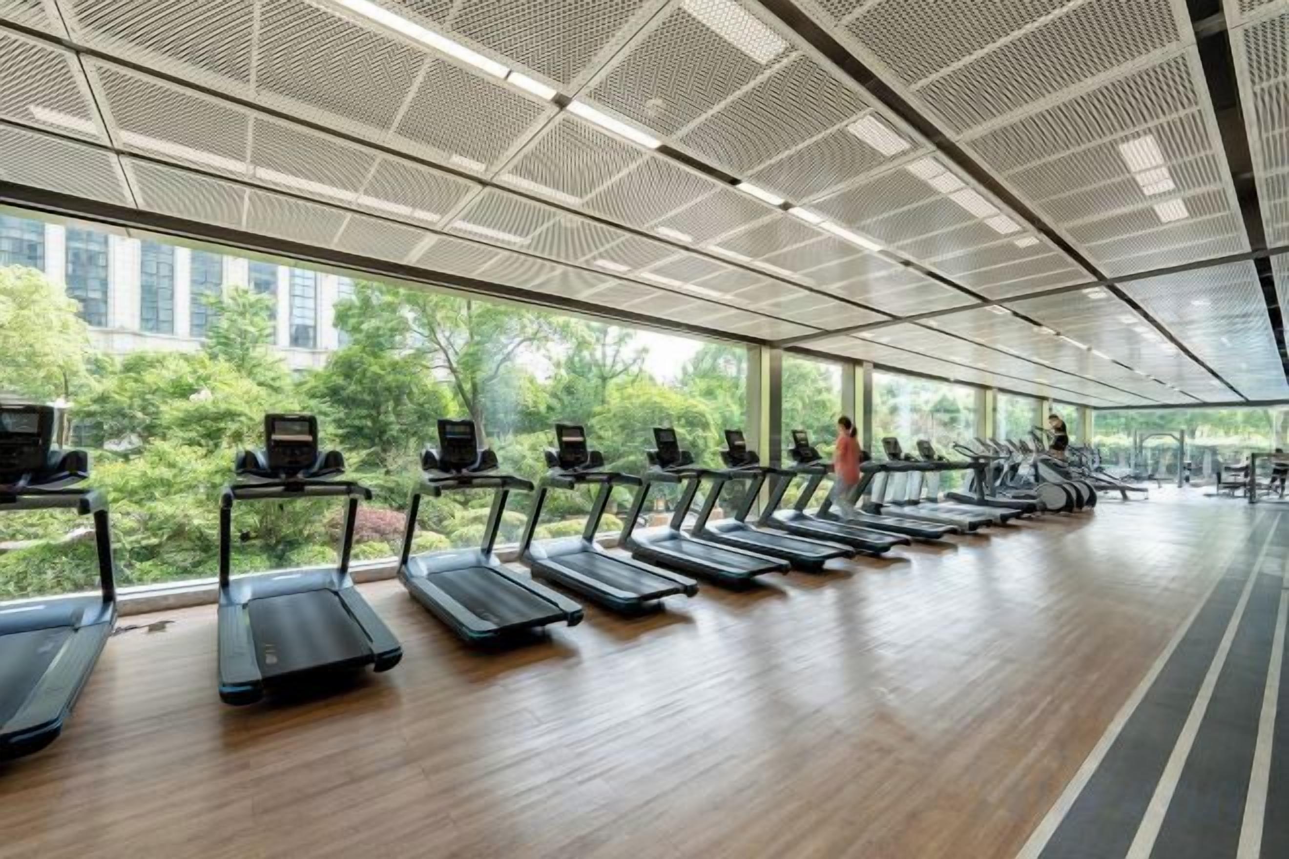 Ein Fitnessraum mit Laufbändern an der Wand, großen Fenstern und Blick auf Grünfläche.