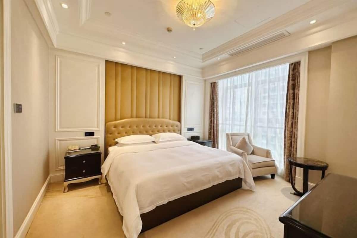 Quarto de hotel com uma cama grande, área de estar com uma poltrona, uma mesa pequena e vista da cidade através de amplas janelas.