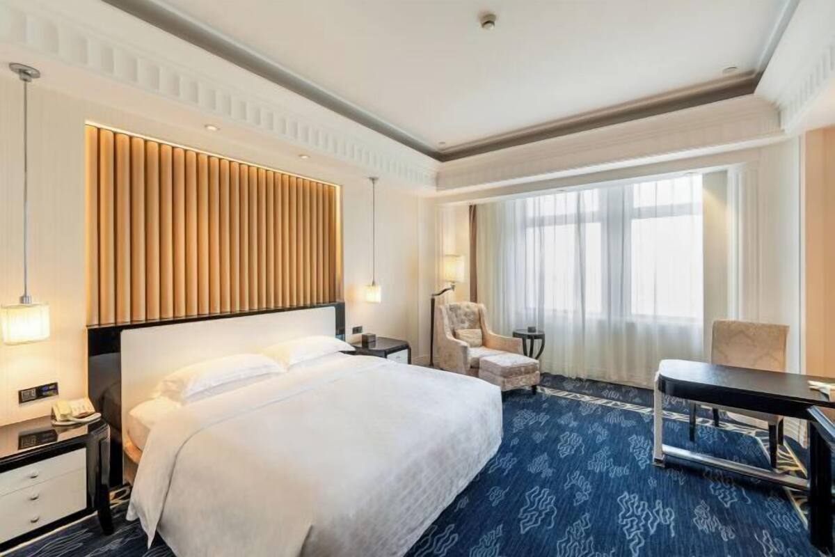 Quarto de hotel com uma cama grande, uma escrivaninha, uma cadeira, uma luminária e uma janela com cortinas.