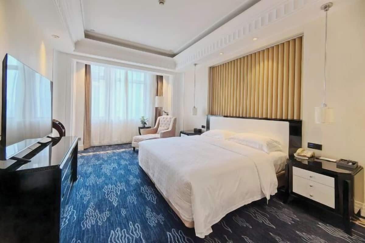 Quarto de hotel com uma cama grande, uma mesa de trabalho com televisão de tela plana, uma cadeira, uma mesa de cabeceira com abajur e uma janela com cortinas.