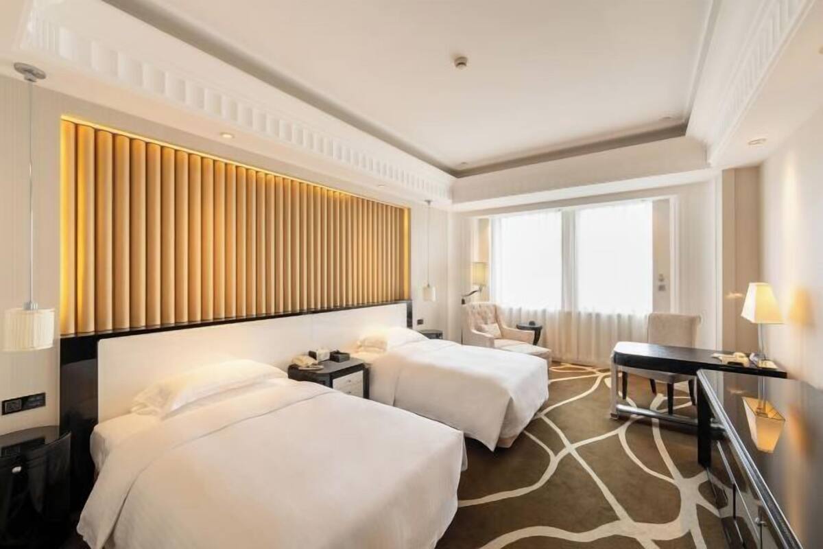 Quarto de hotel com duas camas, uma mesa, uma cadeira e uma janela grande.