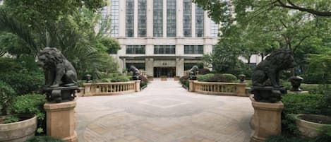 A courtyard with a central paved area, surrounded by greenery and stone lion statues. A tall building with many windows is visible in the background.
