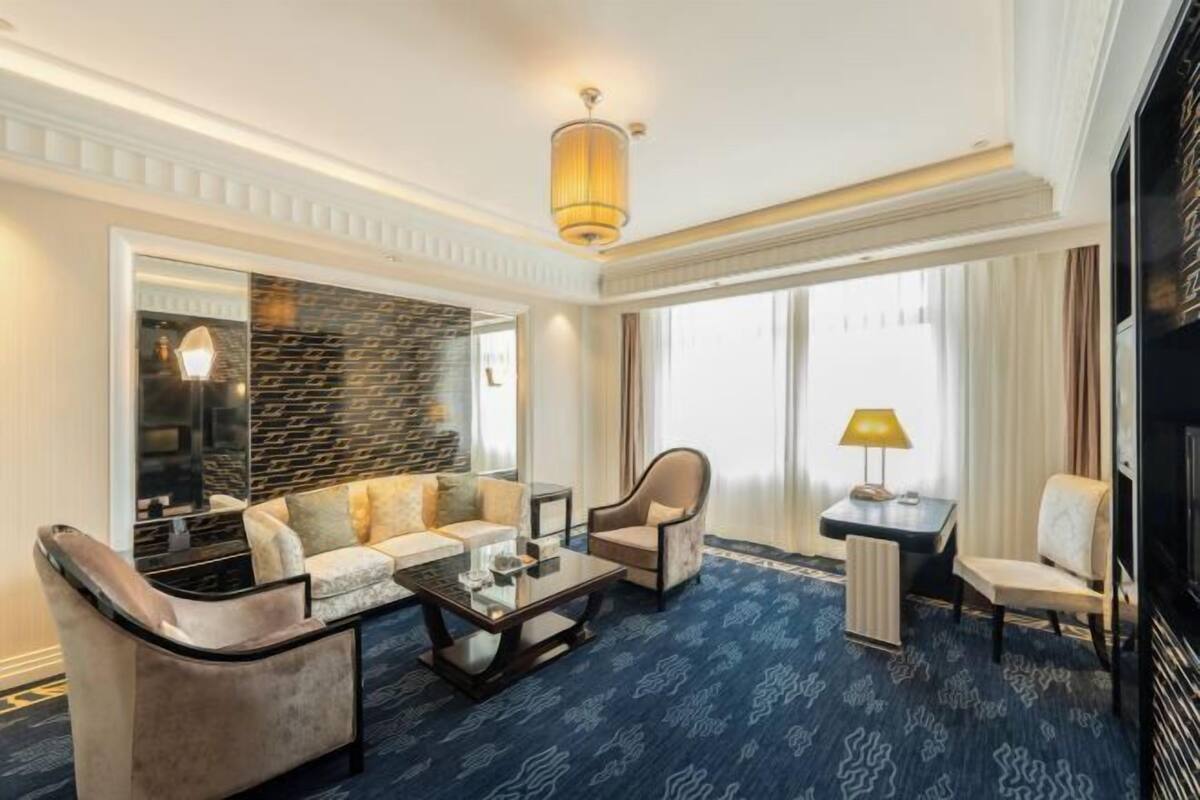 Quarto de hotel com sofá, mesa de centro, abajur e uma grande janela com cortinas.
