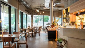 Un café amplio con mesas y sillas de madera, una zona de bar con estanterías llenas de botellas y ventanas grandes que ofrecen vistas al exterior.