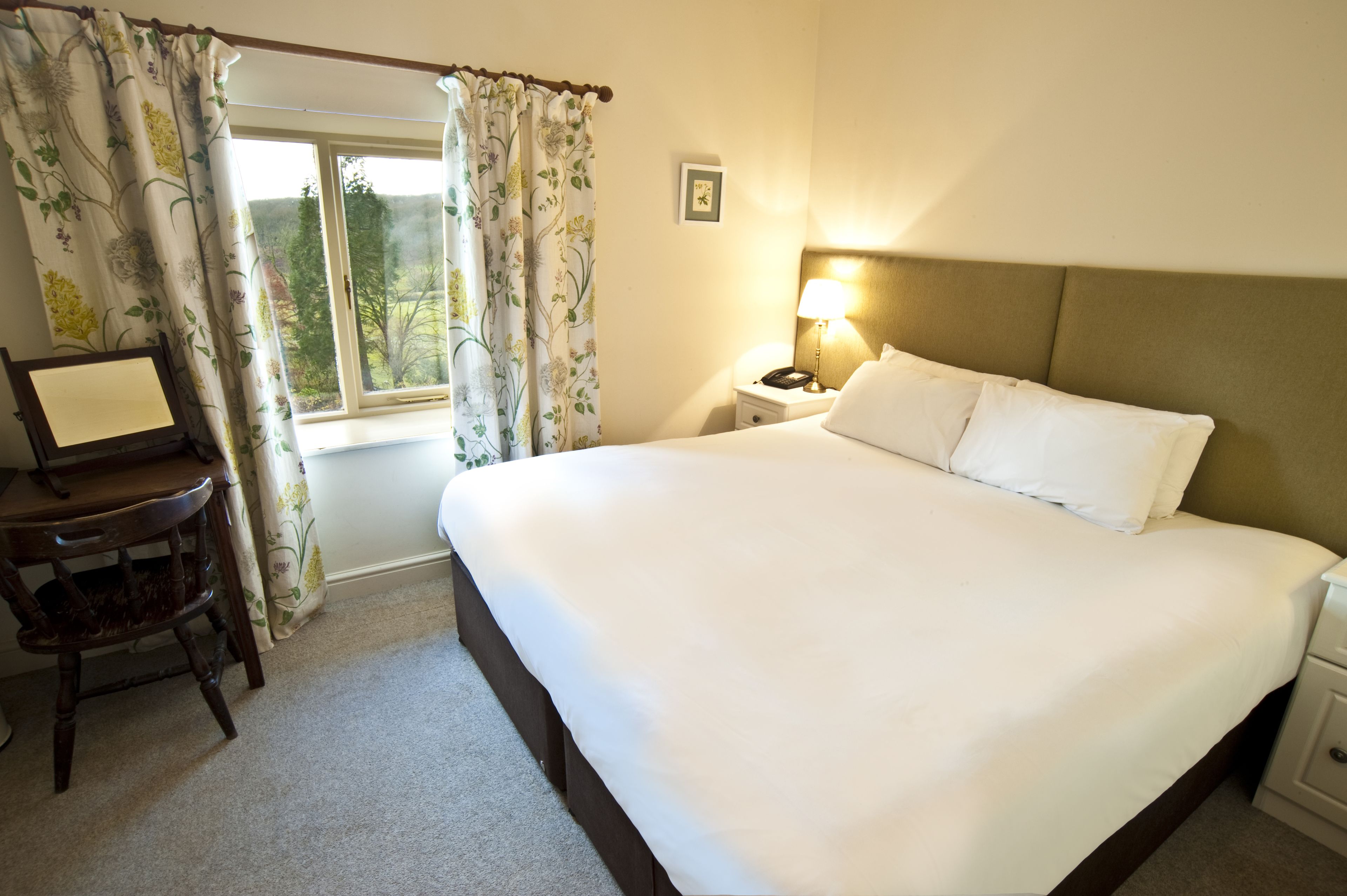 A hotel room with a large bed, a bedside table with a lamp, a chair, and a desk with a computer.