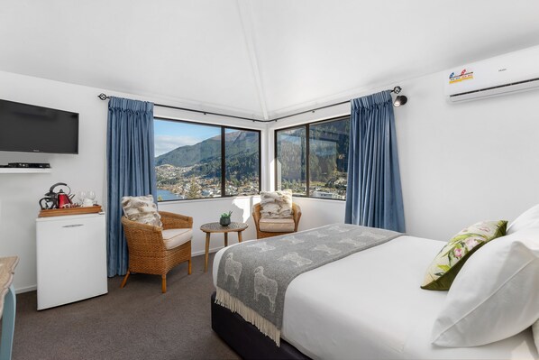 A hotel room with a bed, a TV, a small fridge, two chairs, and a view of mountains. - Balmoral Lodge (Queenstown)