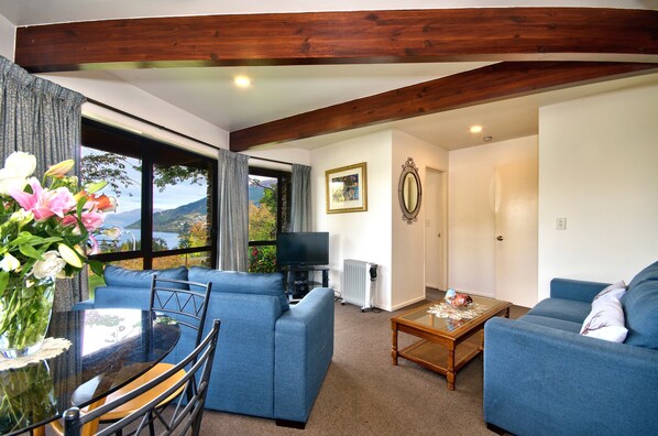 A living room with blue sofas, a glass coffee table, and a TV. There's a window with a view of the outdoors. - Balmoral Lodge (Queenstown)