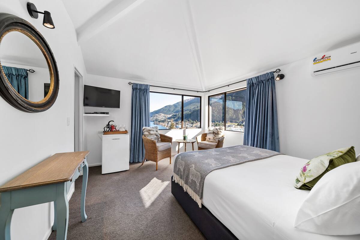 A modern hotel room with a large bed, a desk, a chair, and a TV. The room has a large window with a mountain view.