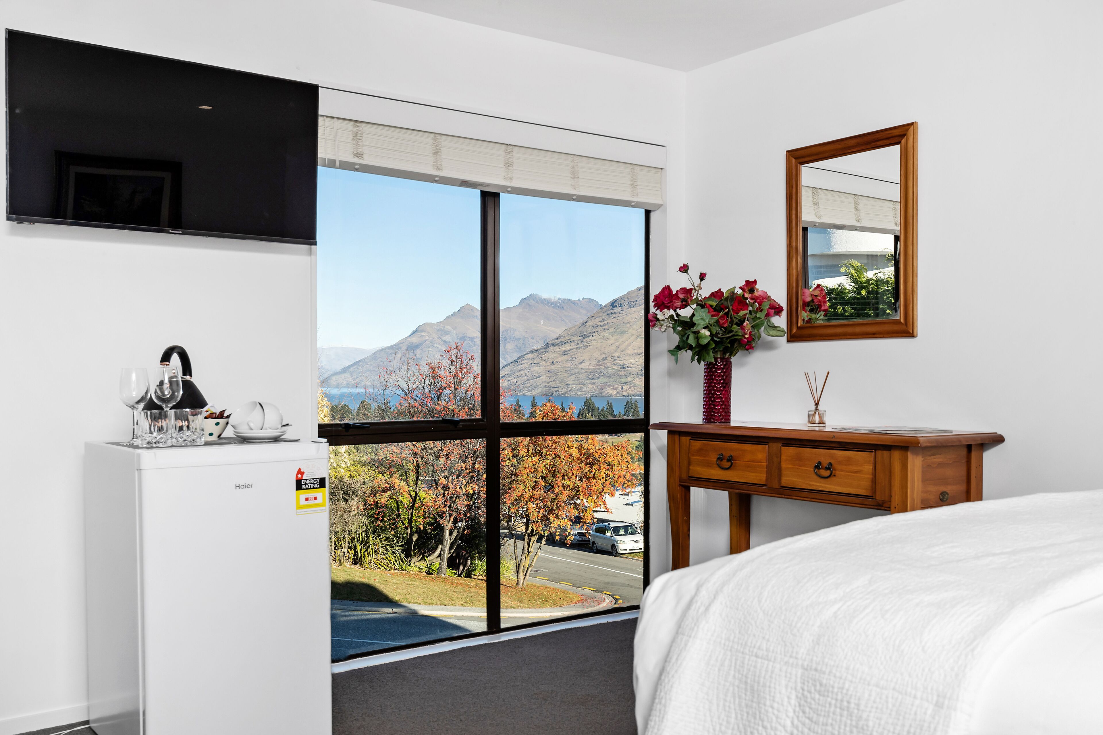A hotel room with a large window offering a mountain view, a wooden dresser, a bed, and a flat-screen TV mounted on the wall.