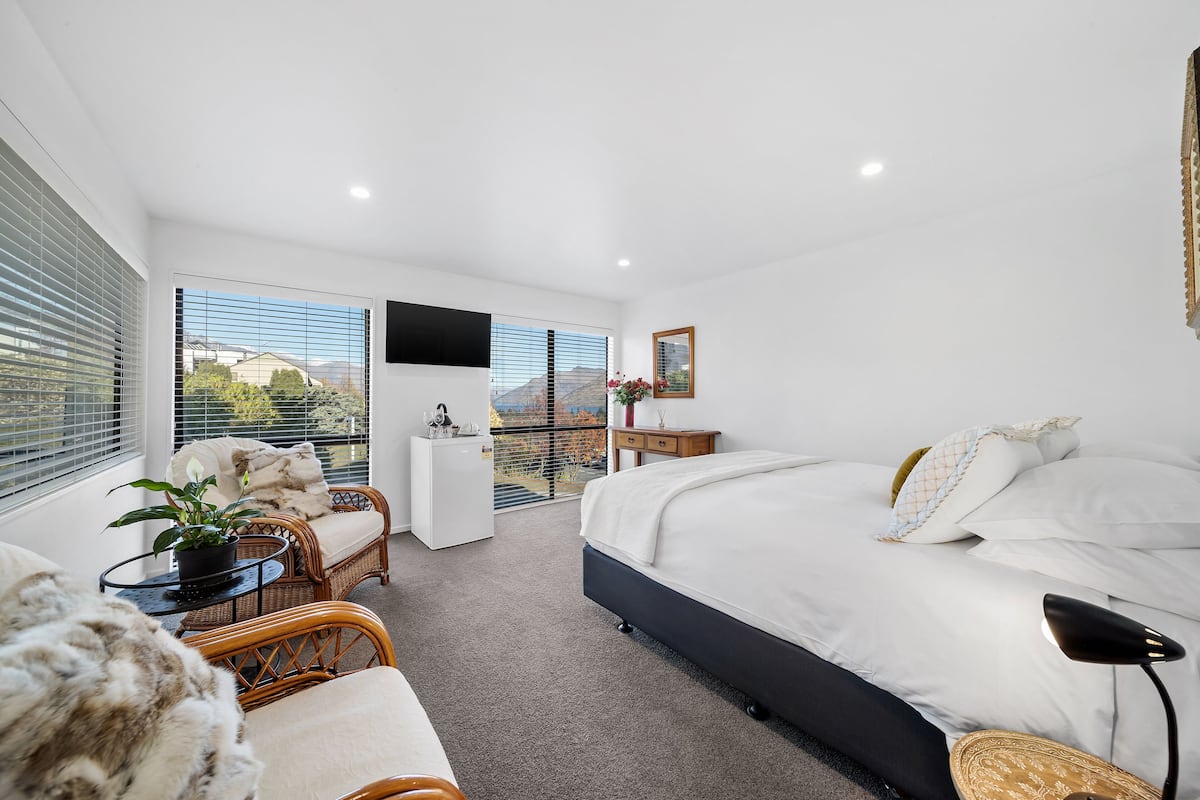 A modern bedroom with a large bed, a seating area with wicker chairs, a small table with a plant, and a balcony with a view.