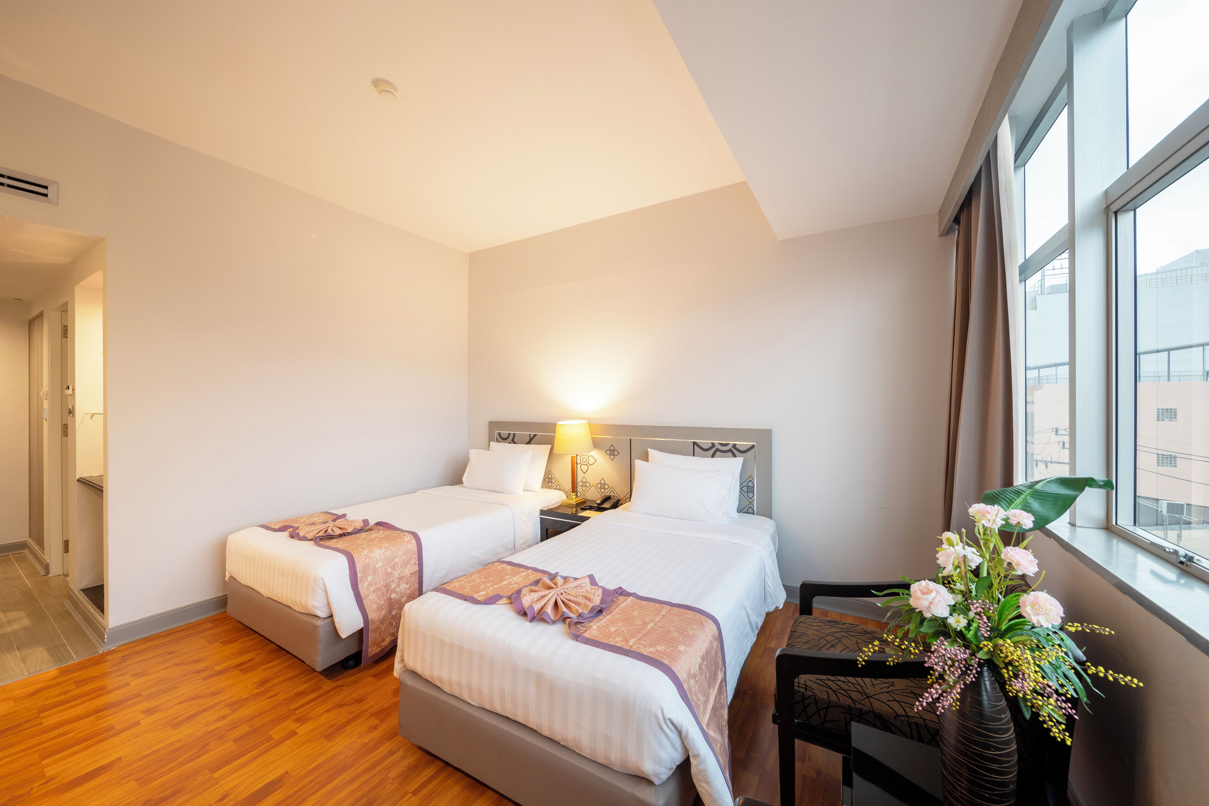 A hotel room with two beds, a nightstand, a desk, and a window with a view of buildings.