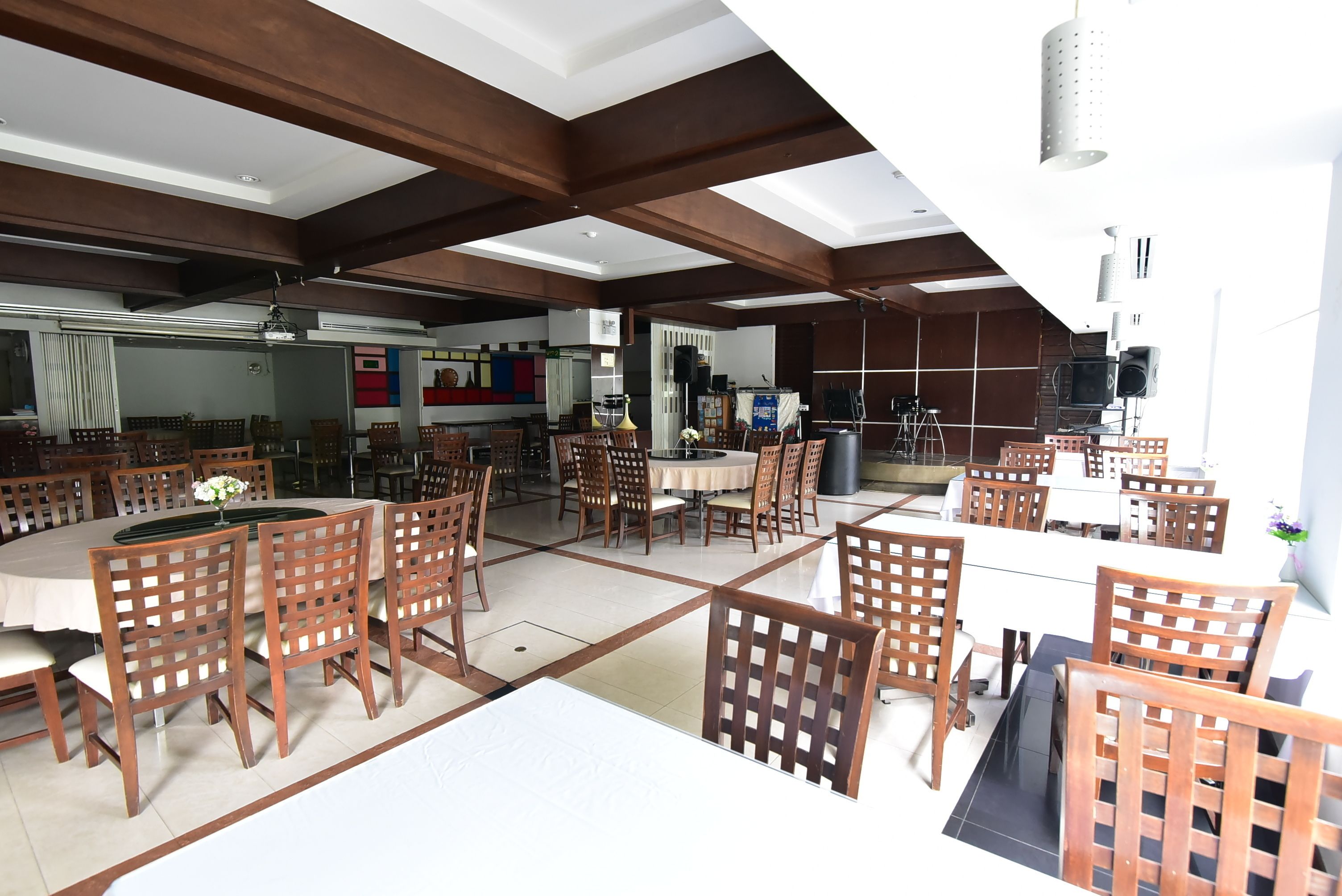 A spacious dining area with multiple tables and chairs, a kitchen in the background, and a modern ceiling design.