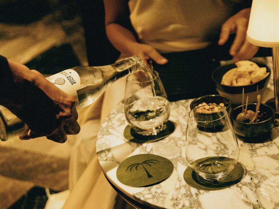 Una mesa con una botella vertiéndose en una copa, un bol de aperitivos y una vela encendida.