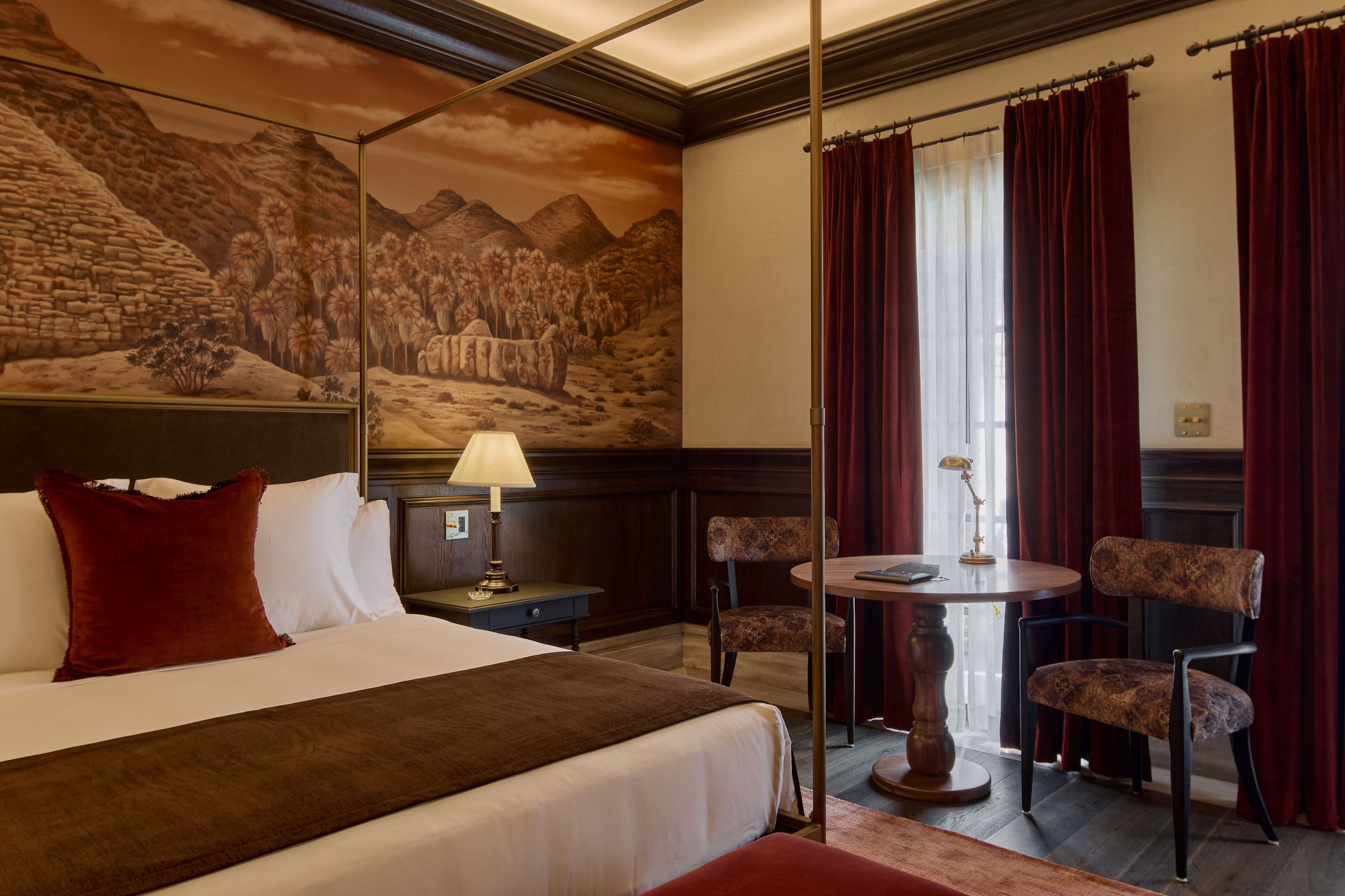 Habitación de hotel con cama, mesita de noche, lámpara, dos sillas, una pequeña mesa y un mural en la pared.