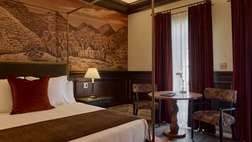 Habitación de hotel con cama, mesita de noche, lámpara, dos sillas, una pequeña mesa y un mural en la pared.