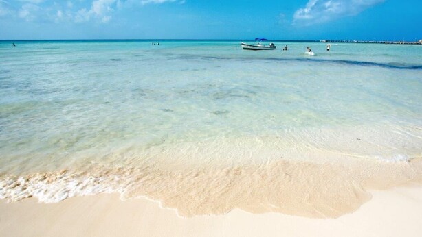 Una playa con agua turquesa cristalina, una orilla arenosa y un barco a lo lejos.